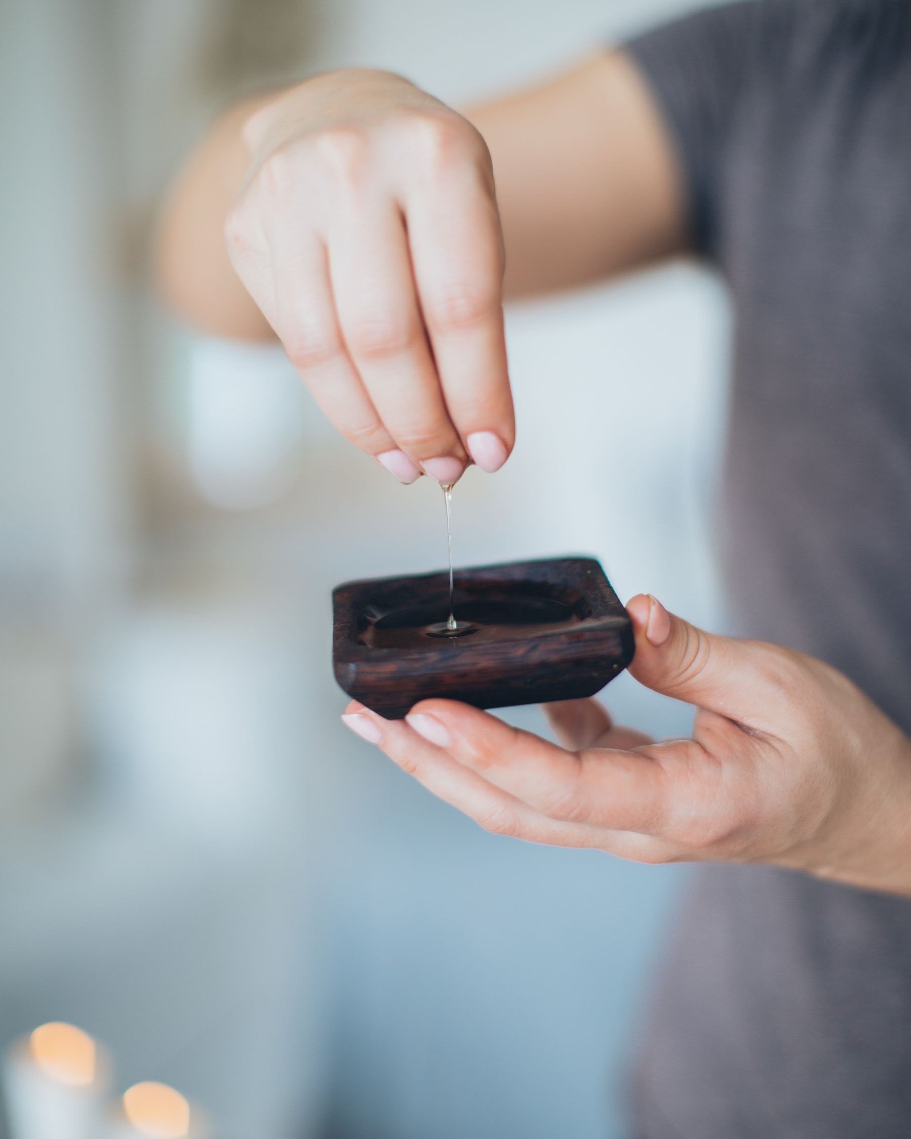 Manos de una esteticista dejando caer unas gotas de aceite en un cuenco de madera oscura, con velas desenfocadas al fondo